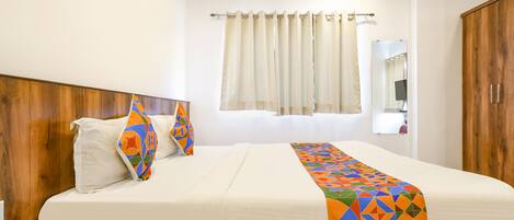 A hotel room with a wooden headboard, a bed with white bedding, a colorful geometric throw blanket, a bedside table with a phone, and a window with curtains.