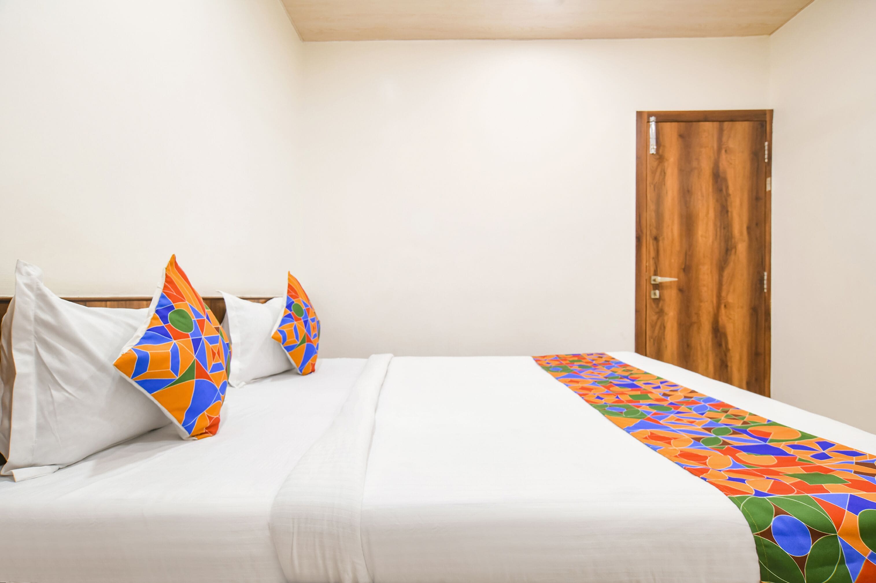 Habitación de hotel con cama, ropa de cama blanca, almohadas coloridas, una puerta de madera y un corredor de cama estampado.