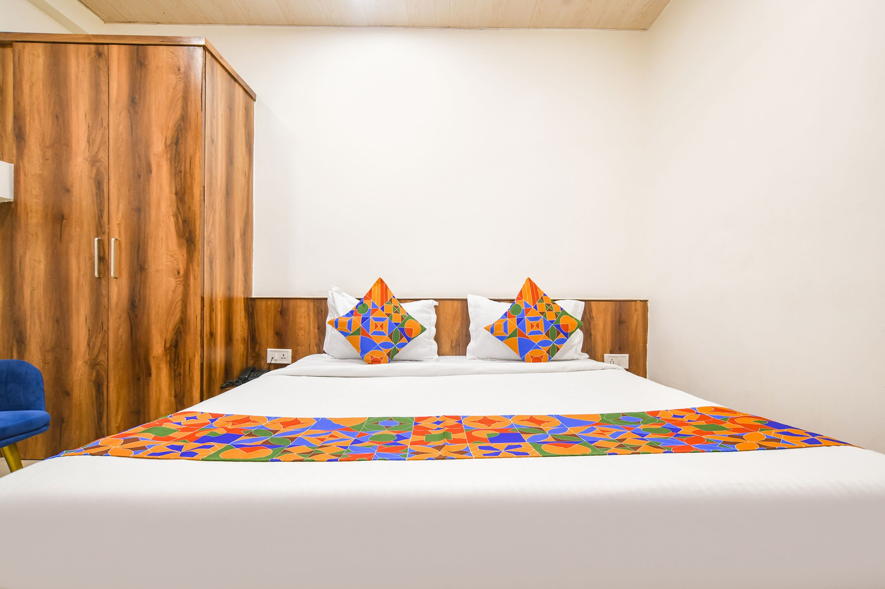 A hotel room with a wooden wardrobe, a bed with white bedding, two decorative pillows, and a colorful geometric-patterned bedspread.