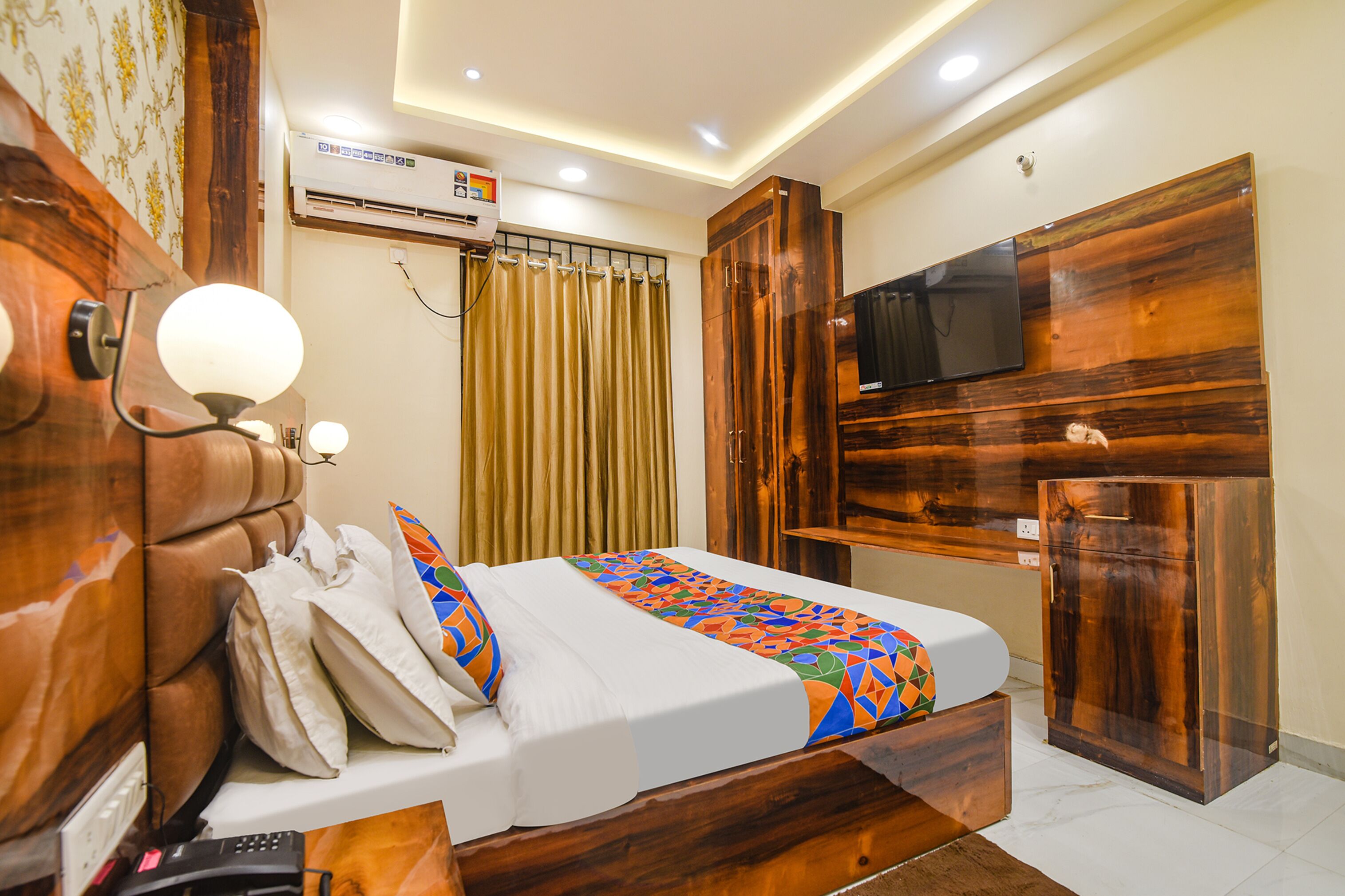 A hotel room with a wooden headboard, a bed with a colorful geometric-patterned bedspread, a flat-screen TV mounted on a wooden cabinet, and an air conditioning unit on the wall.
