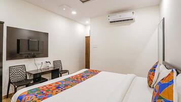 Et hotelværelse med seng, fjernsyn ophængt på væggen, skrivebord med stole og en aircondition.