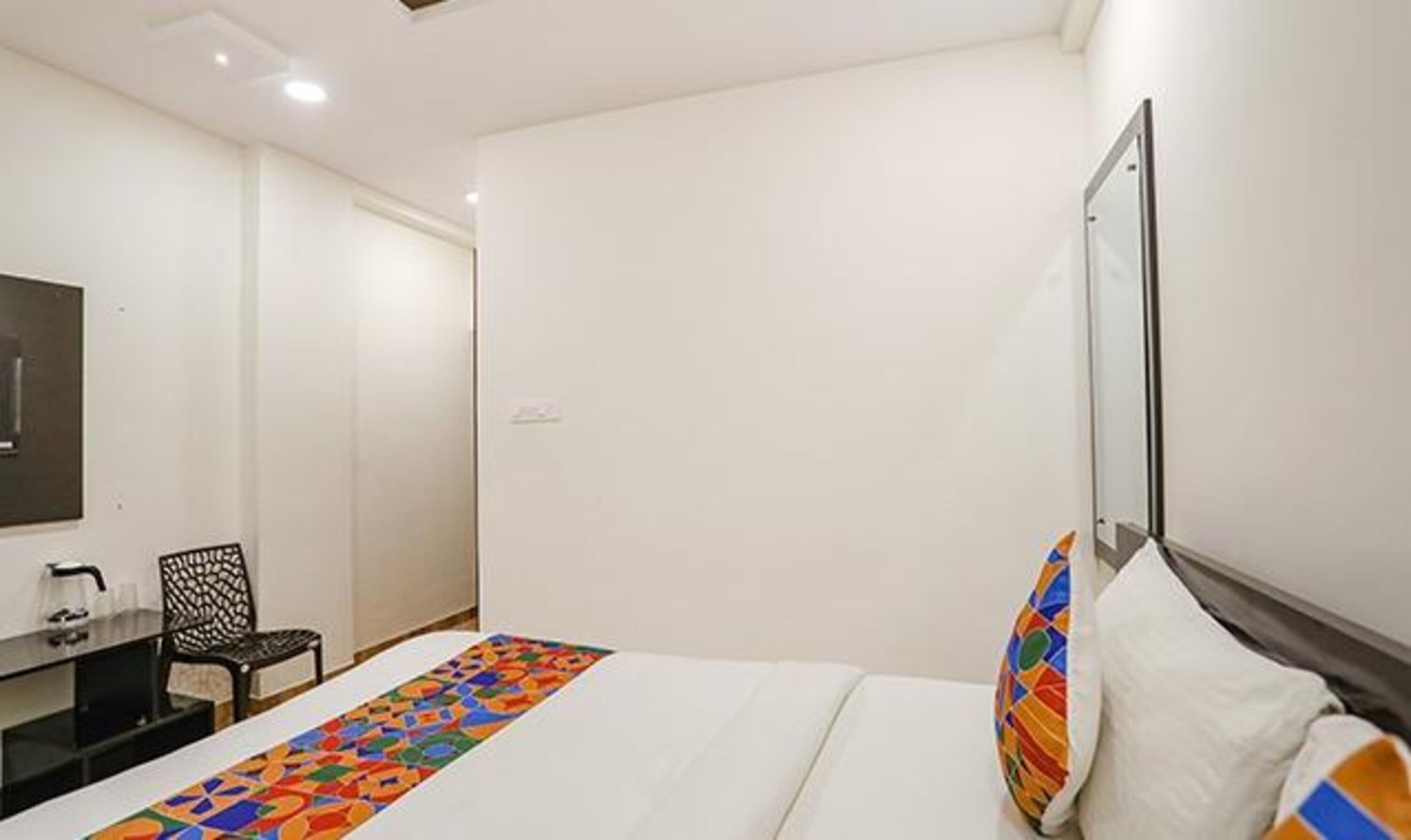 A hotel room with a bed, a desk with a chair, a mirror, and a colorful rug.