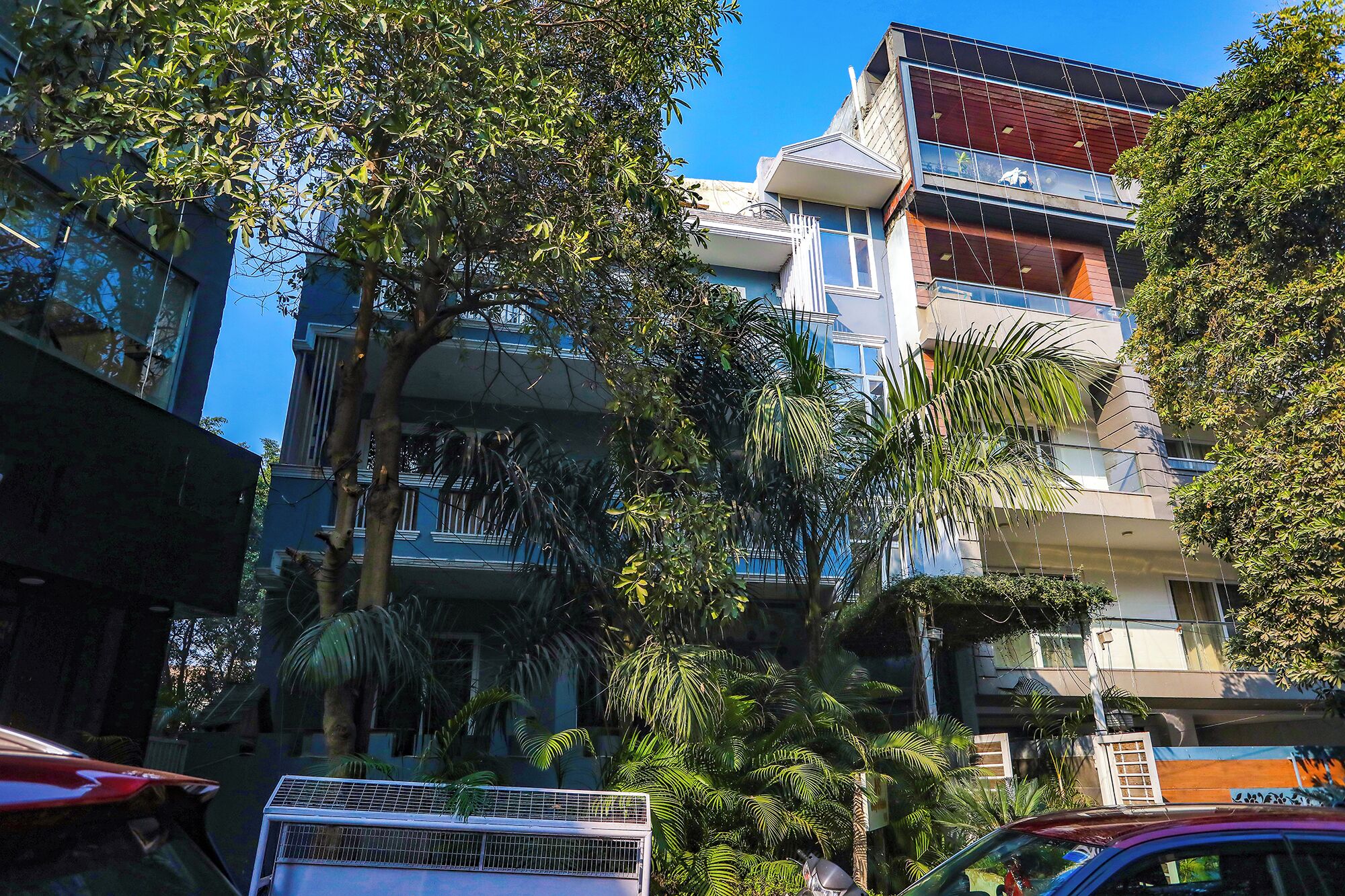 A multi-story building with balconies, surrounded by trees and parked cars.