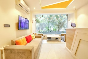 A modern living room with a sofa, a coffee table, a TV, and a large window with a view of greenery. - FabHotel JHS Studio- Nr. Phase 1 Metro Station Gurgaon (Gurugram)