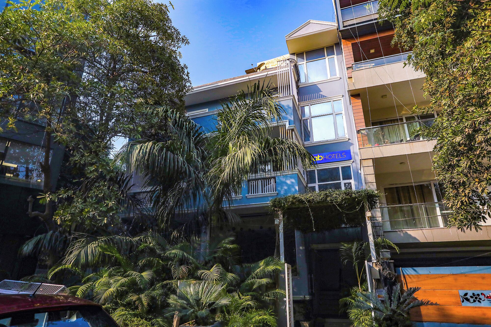 Un edificio de varios pisos con balcones, un cartel que dice 'ab+HOTELS' y abundante vegetación circundante.