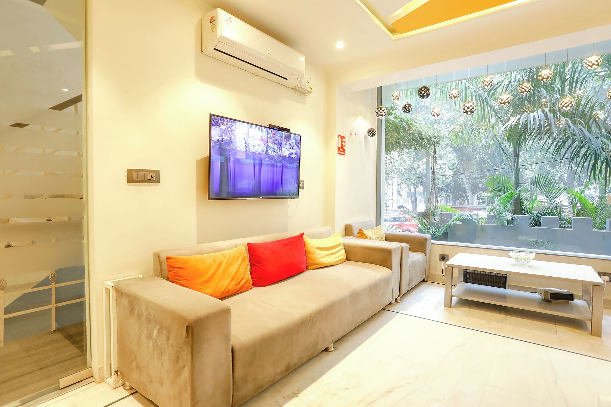 A modern living room with a beige sofa, a flat-screen TV, and a large window with a view of greenery.