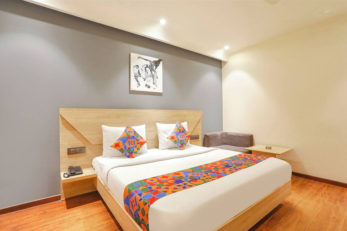 Habitación de hotel con cabecera de madera, cama con ropa blanca, dos almohadas geométricas coloridas, una mesita de noche con un teléfono y un control remoto, y un pequeño sofá gris.