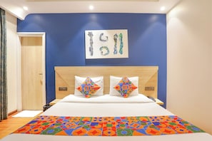 A bedroom with a blue wall, a bed with white bedding, two pillows with colorful patterns, and a framed artwork above the bed. - FabHotel JHS Studio- Nr. Phase 1 Metro Station Gurgaon (Gurugram)