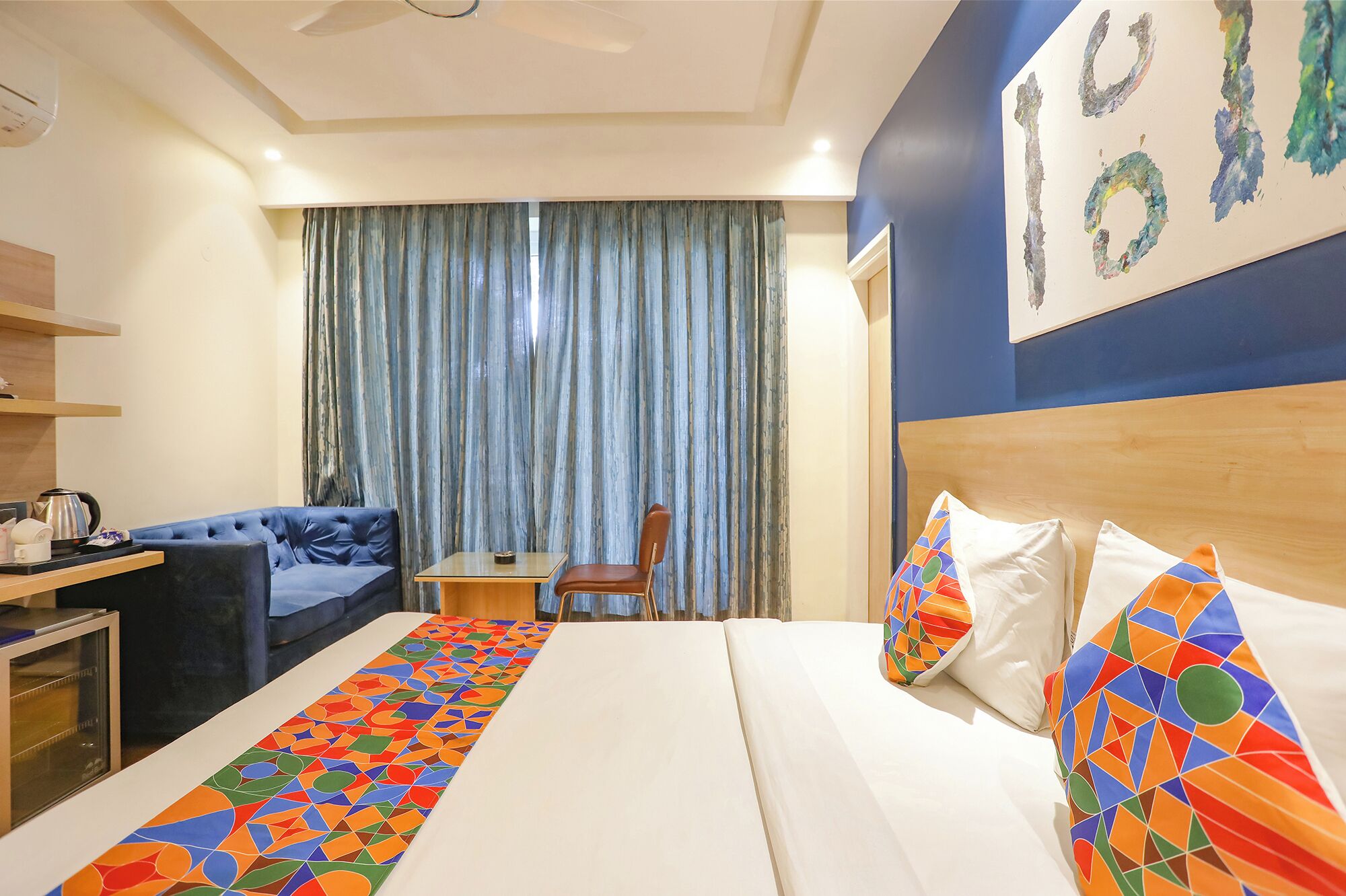 A hotel room with a bed, a desk, a chair, a blue sofa, and a large window with curtains.