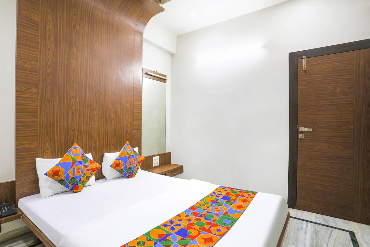 Een hotelkamer met een houten hoofdeinde, een bed met wit beddengoed, kleurrijke kussens en een kamerkleed met een patroon.