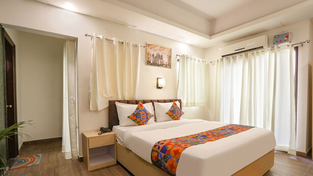 A hotel room with a large bed, a bedside table, a nightstand, a wall-mounted air conditioning unit, and a window with curtains. - FabHotel The Indian (Gurugram)