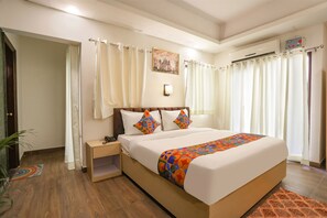 A hotel room with a large bed, a bedside table, a nightstand, a wall-mounted air conditioning unit, and a window with curtains. - FabHotel The Indian (Gurugram)