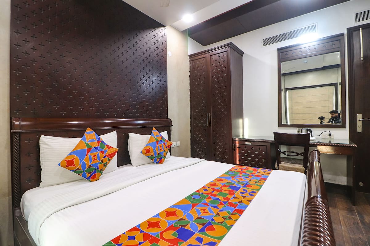 A hotel room with a large bed, two pillows, a wooden headboard, a wooden wardrobe, a small desk with a mirror, and a wooden chair.