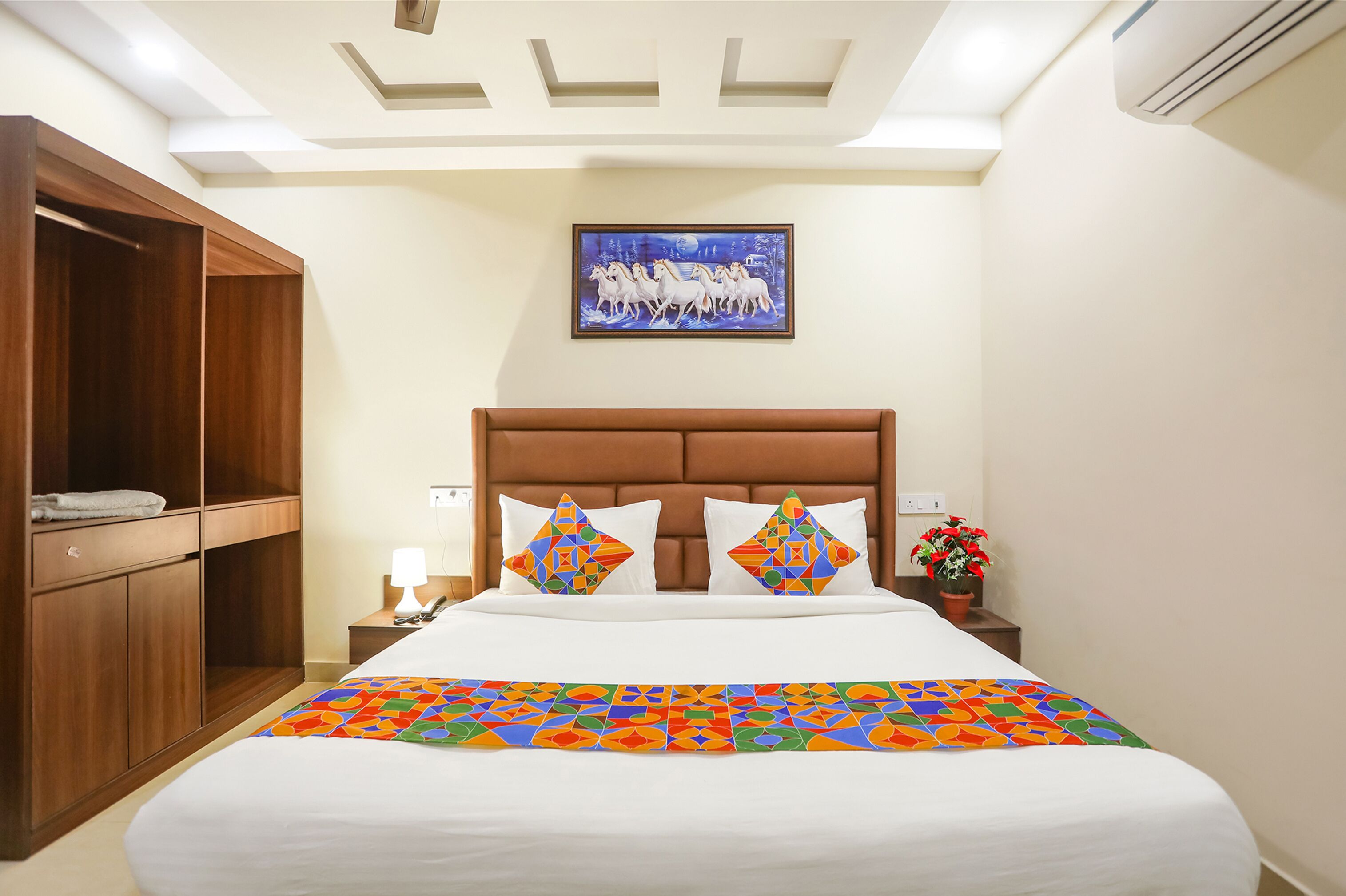 A hotel room with a bed, a nightstand, a wooden wardrobe, a painting on the wall, and a potted plant.