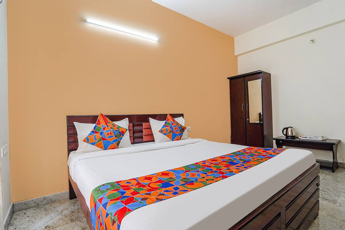 A neatly made bed with a colorful geometric-patterned bedspread, a wooden headboard, and a bedside table with a kettle and a cup.