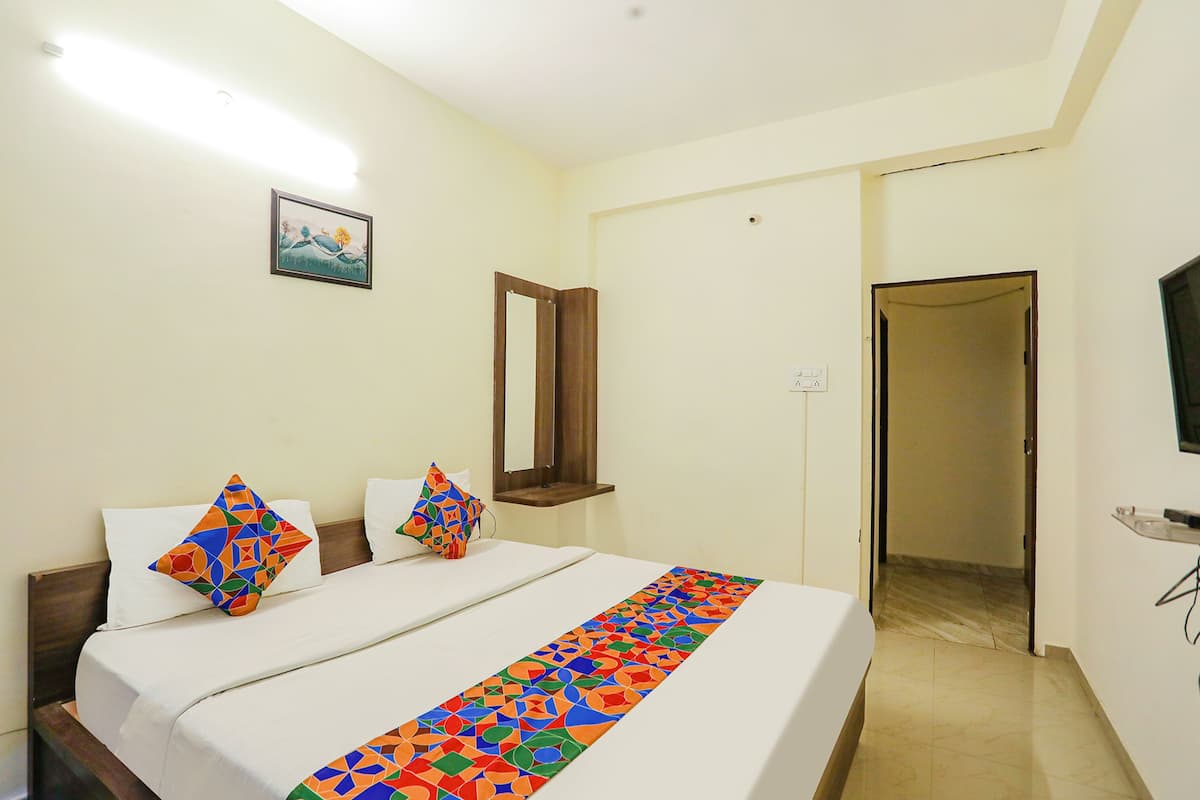 A hotel room with a bed, two pillows, a colorful bed runner, a wall-mounted TV, a painting, and a door leading to another room.