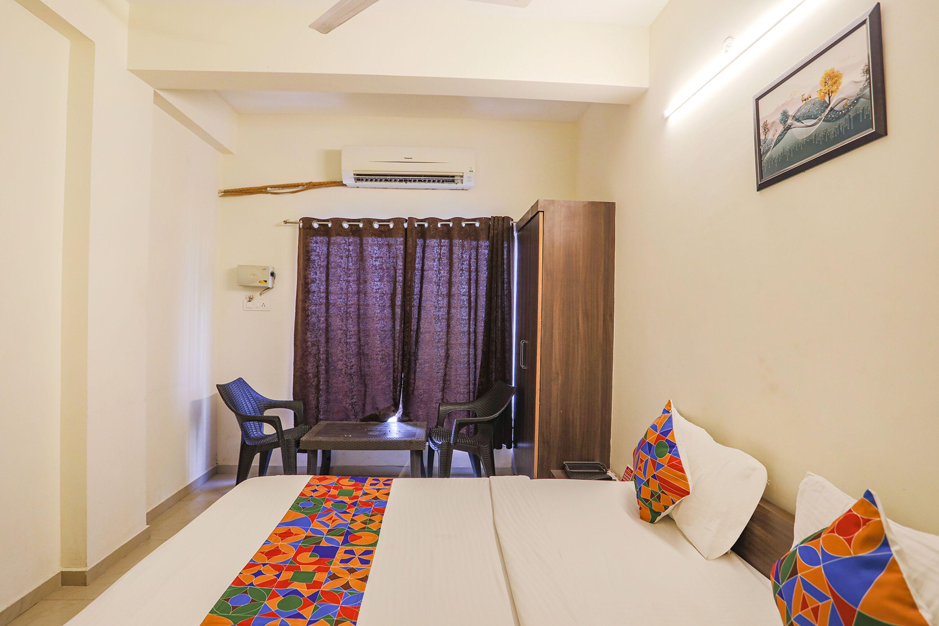 A hotel room with a bed, two chairs, a table, an air conditioning unit, a curtain, and a painting on the wall.