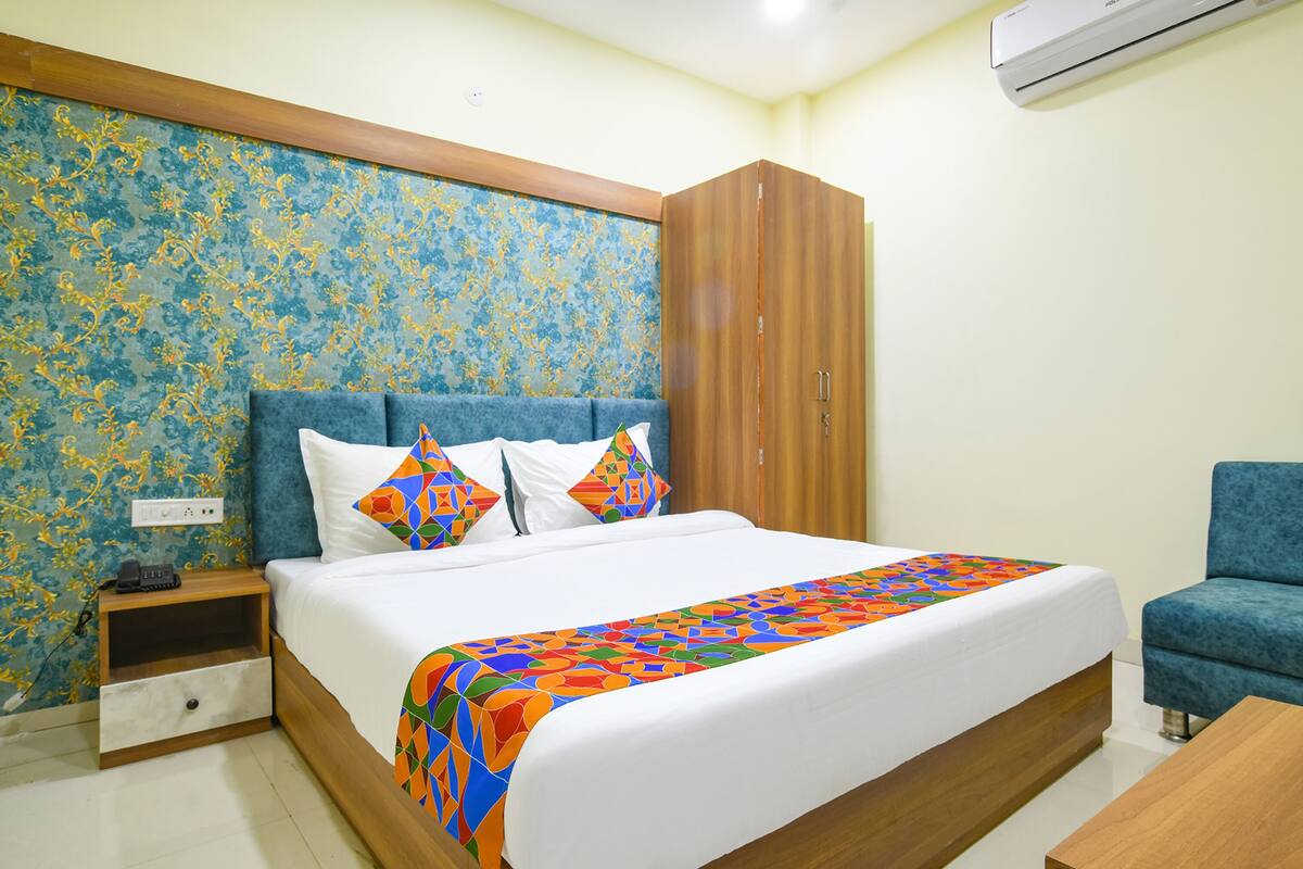 A neatly made bed with a colorful blanket, a wooden wardrobe, a small bedside table with a phone, and a blue armchair.