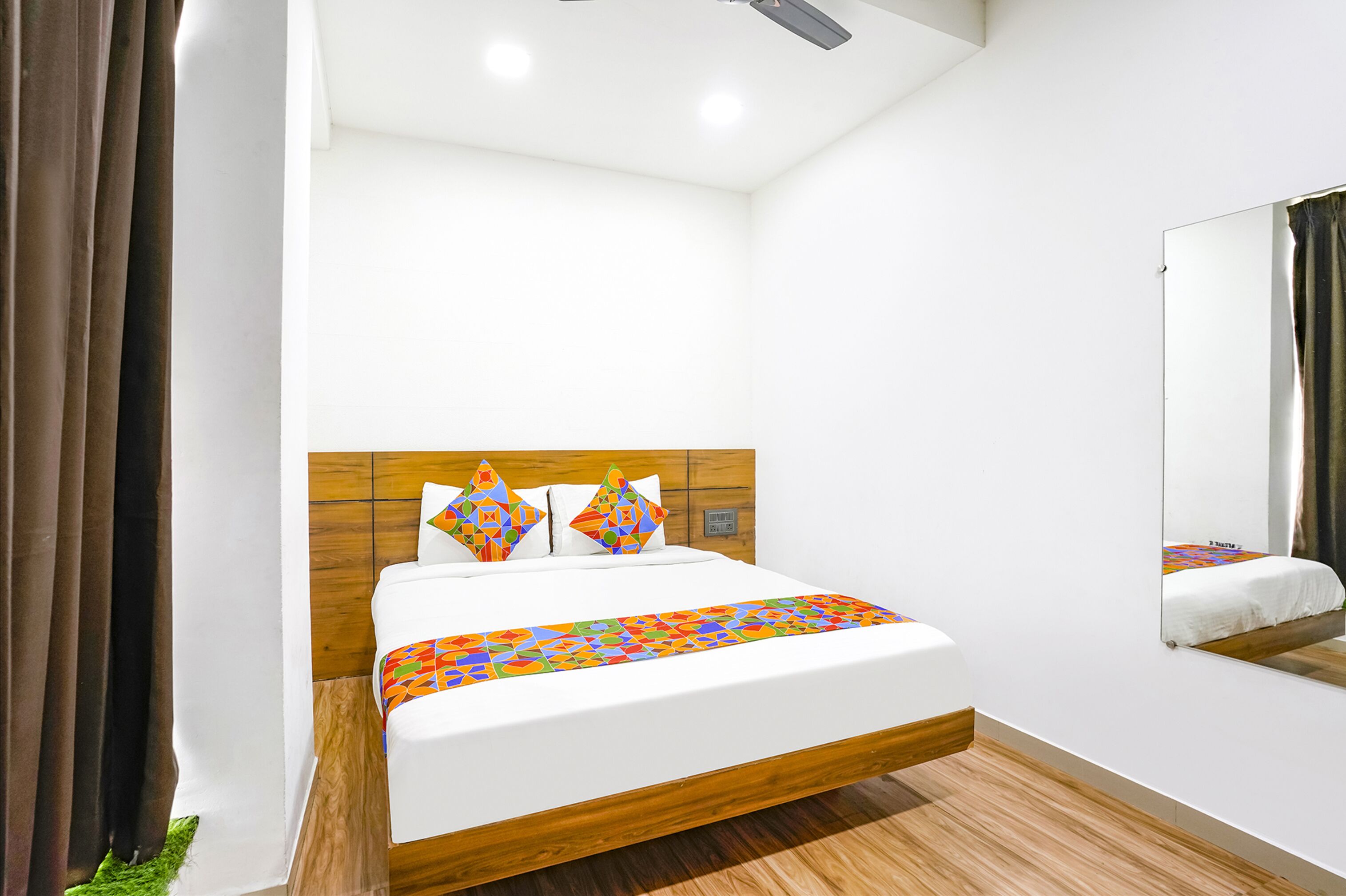 A modern hotel room with a wooden headboard, a large bed with colorful pillows, and a mirror on the wall.