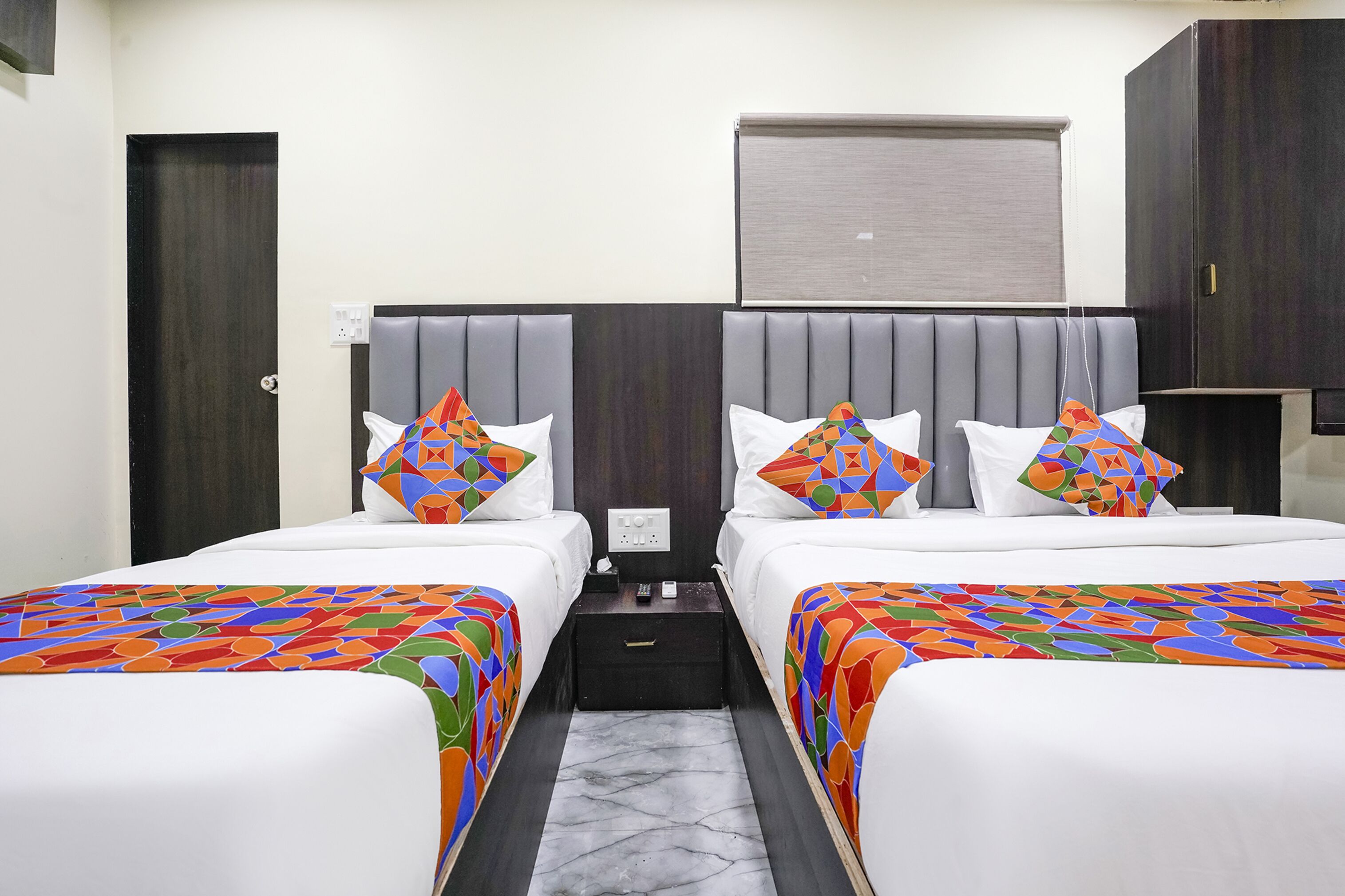 A hotel room with two beds, each with a colorful bedspread and matching pillows. There is a small bedside table with a lamp and a power outlet.
