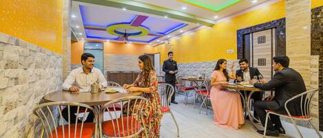 A restaurant with multiple tables and chairs, a waiter, and a ceiling with colorful lighting.