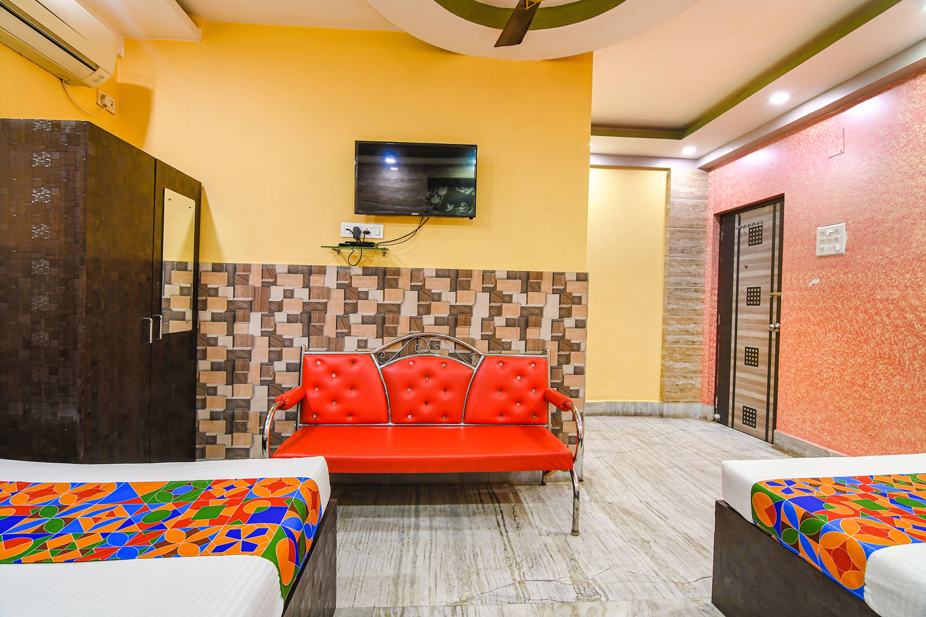 Habitación de hotel con un sofá rojo, dos camas con ropa de cama colorida, un televisor montado en la pared y un papel tapiz estampado.