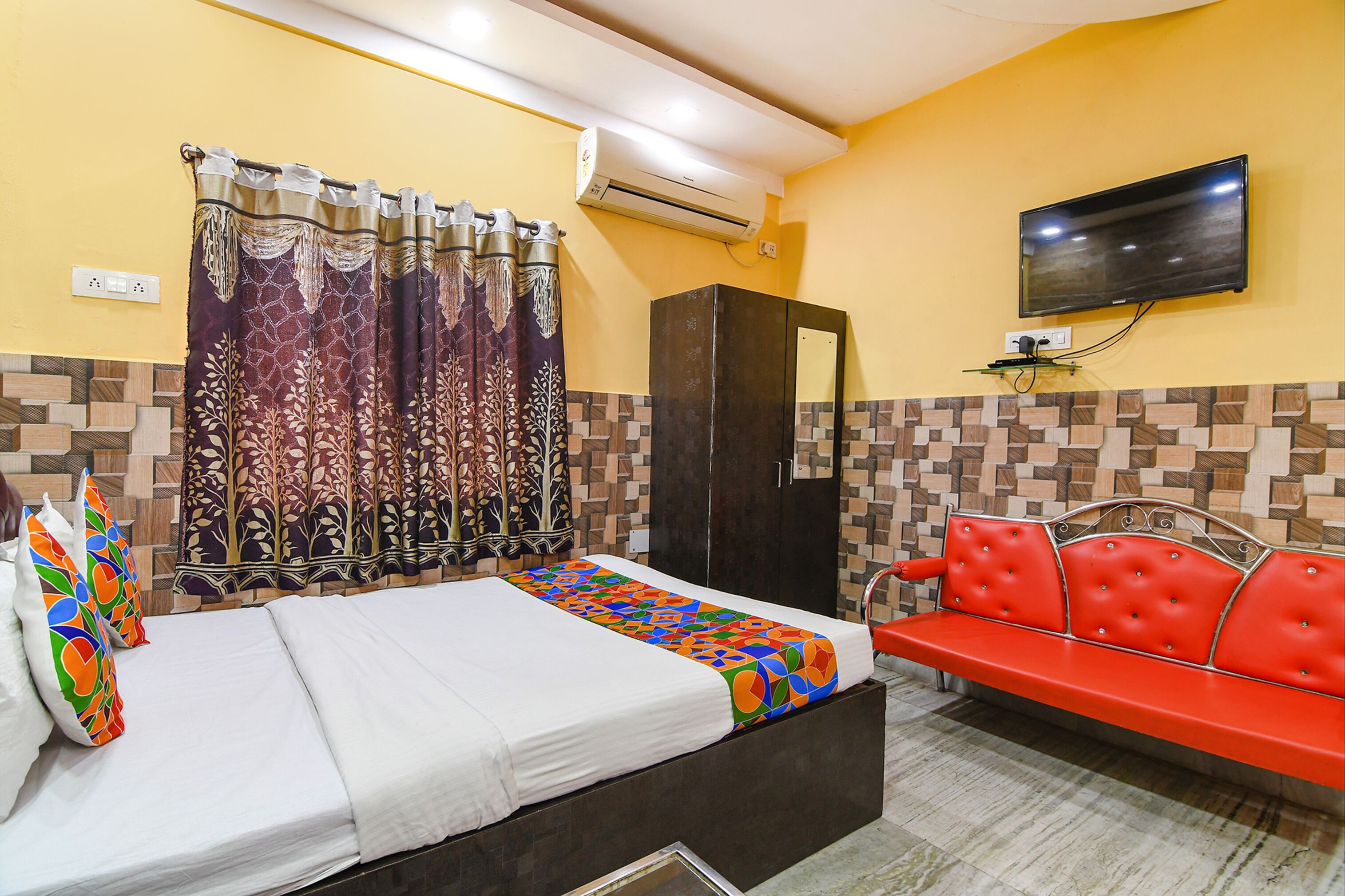 A hotel room with a bed, a bench, a TV, and an air conditioning unit.