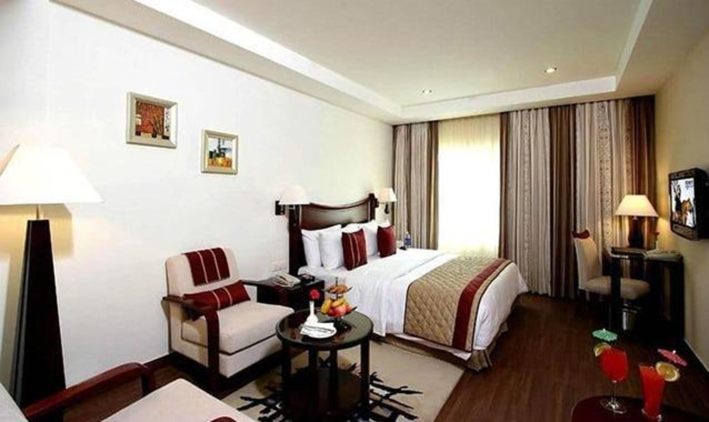 A hotel room with a bed, a chair, a small table with a fruit bowl, a TV, and a lamp.