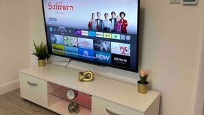 A flat-screen TV on a white entertainment unit with a digital display, a small plant, and a decorative item. - 3-bed Home, Sleeps 8, Ideal for Large Groups (Birmingham)