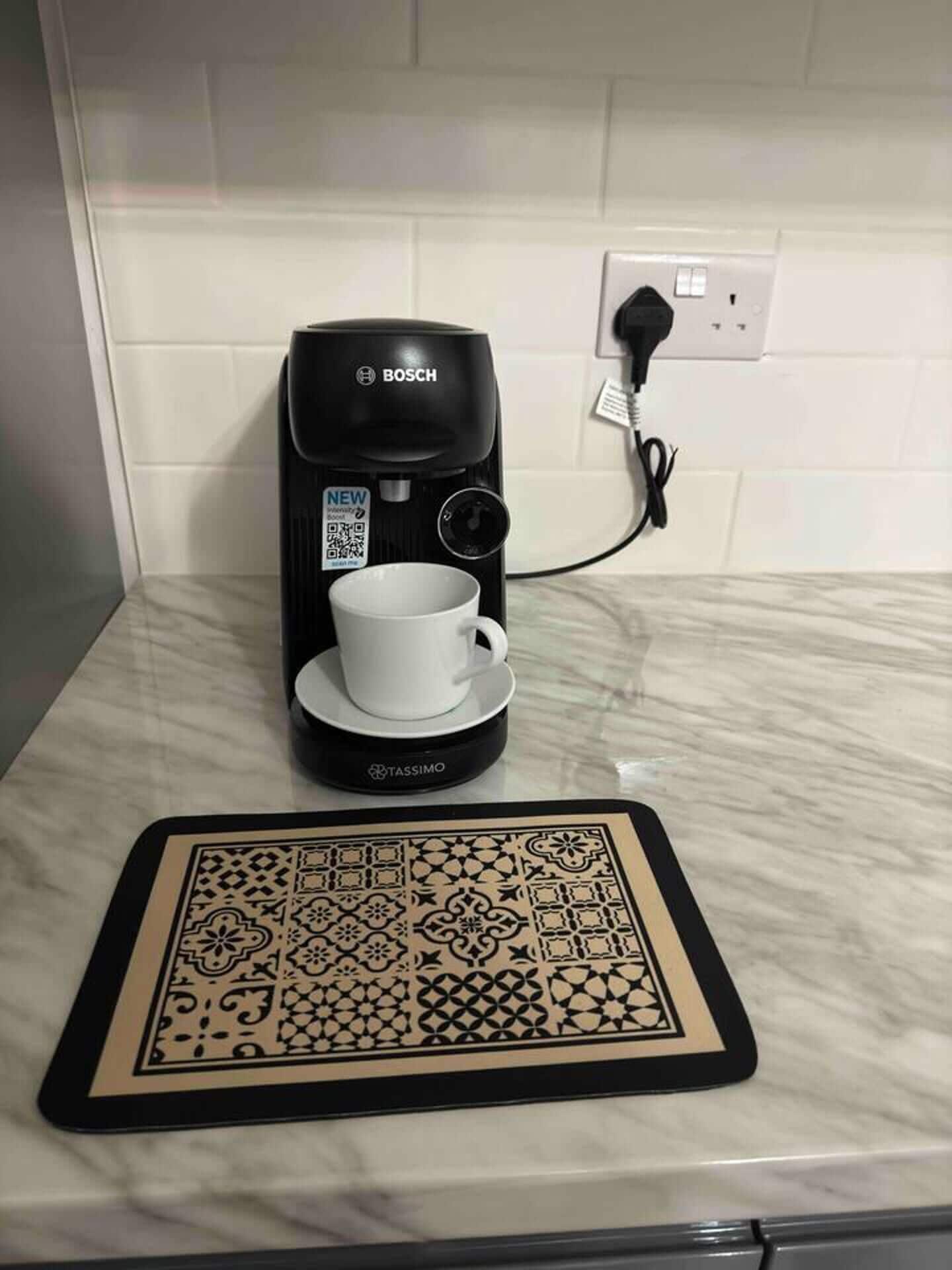 Una cafetera Bosch con una cápsula Tassimo, una taza blanca y un posavasos decorativo sobre una encimera de mármol.