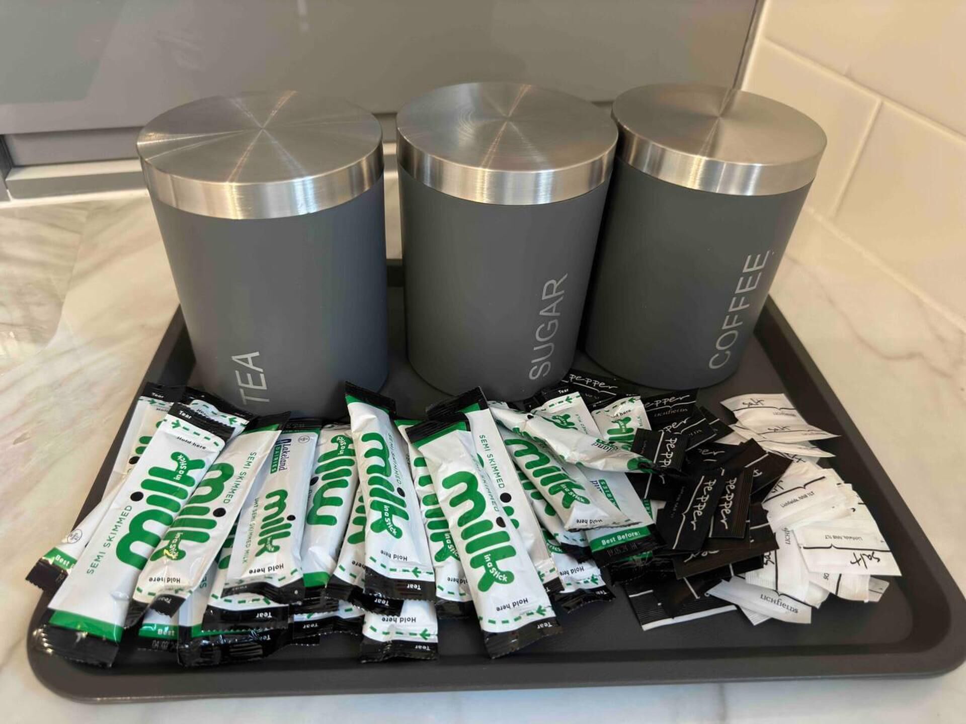 A tray with three metallic containers labeled 'TEA', 'SUGAR', and 'COFFEE', and several packets of 'milk' and 'honey' on a countertop.