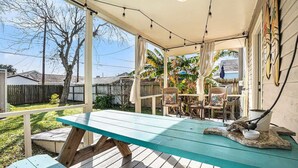 Un patio techado al aire libre con mesa y sillas de madera, una parrilla y vistas al jardín trasero y al vecindario.