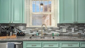 Una cocina con armarios verde azulado, encimera de granito y ventana con persianas.