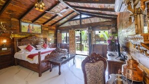 A wooden room with a bed, a table, and a chair. There is a painting on the wall and a window with a view of greenery. - Little Cove Nature Resort (Nashik)