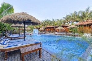A pool area with lounge chairs and thatched umbrellas, surrounded by palm trees and wooden huts. - Little Cove Nature Resort (Nashik)
