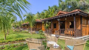 A wooden bungalow with a porch, surrounded by lush greenery and palm trees. - Little Cove Nature Resort (Nashik)