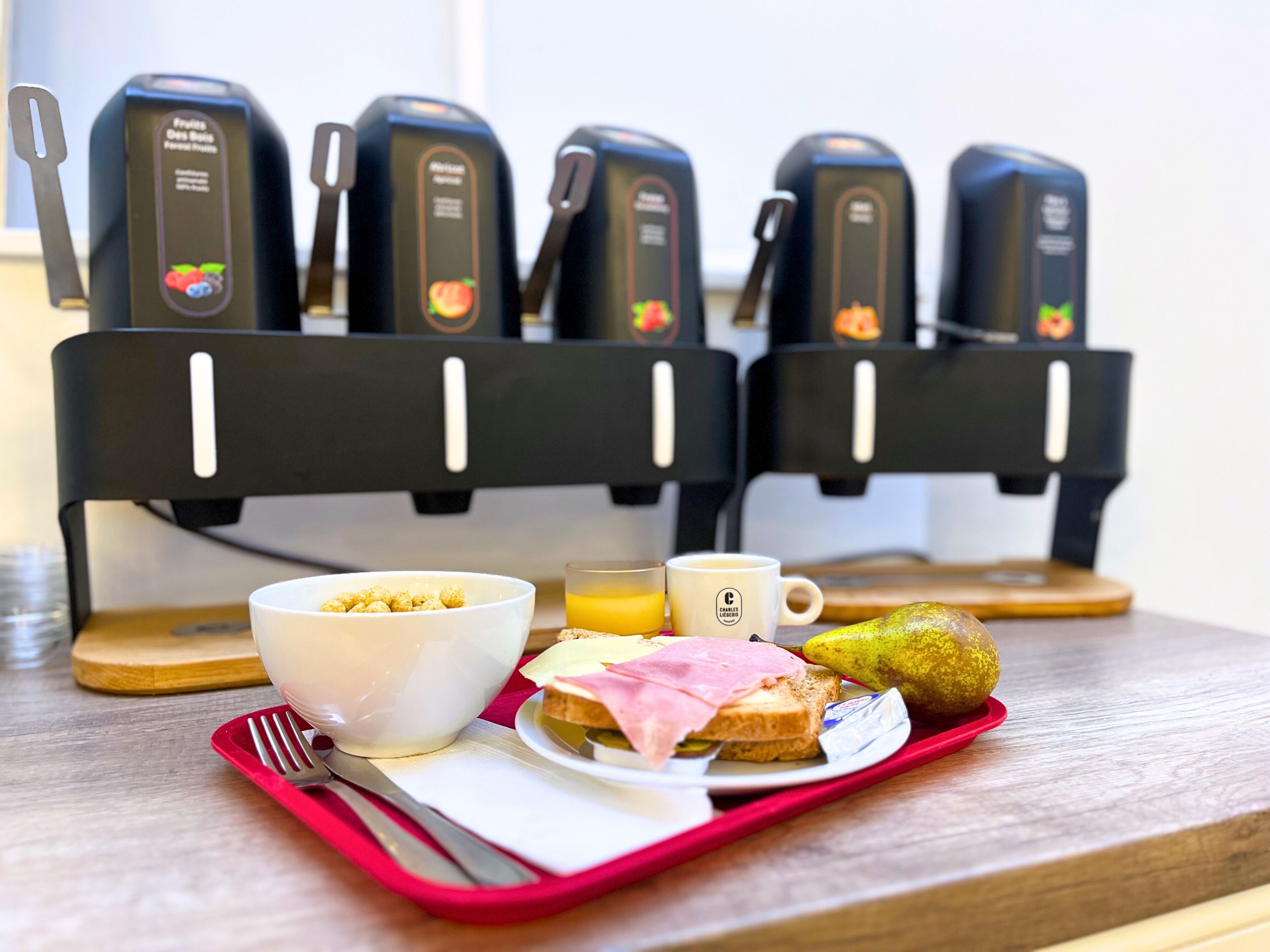 A breakfast tray with cereal, juice, a mug, toast with ham, and a banana on a wooden table with a coffee machine in the background.
