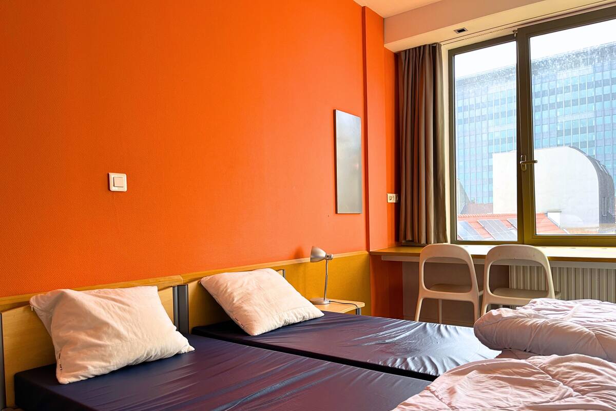 A hotel room with two beds, a desk, a chair, and a window with a view of buildings.