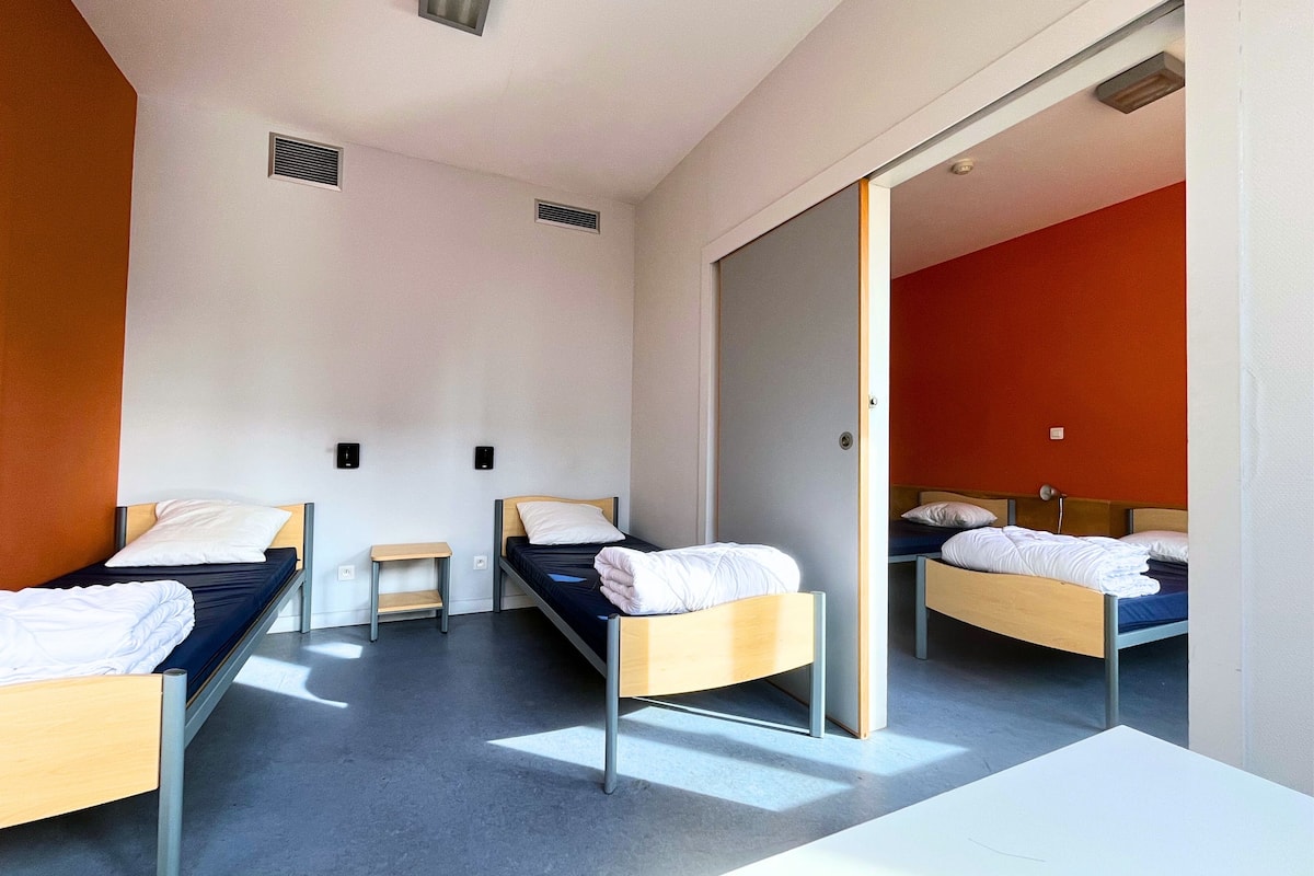 A hotel room with three single beds, a small table, and a sliding door leading to another room.