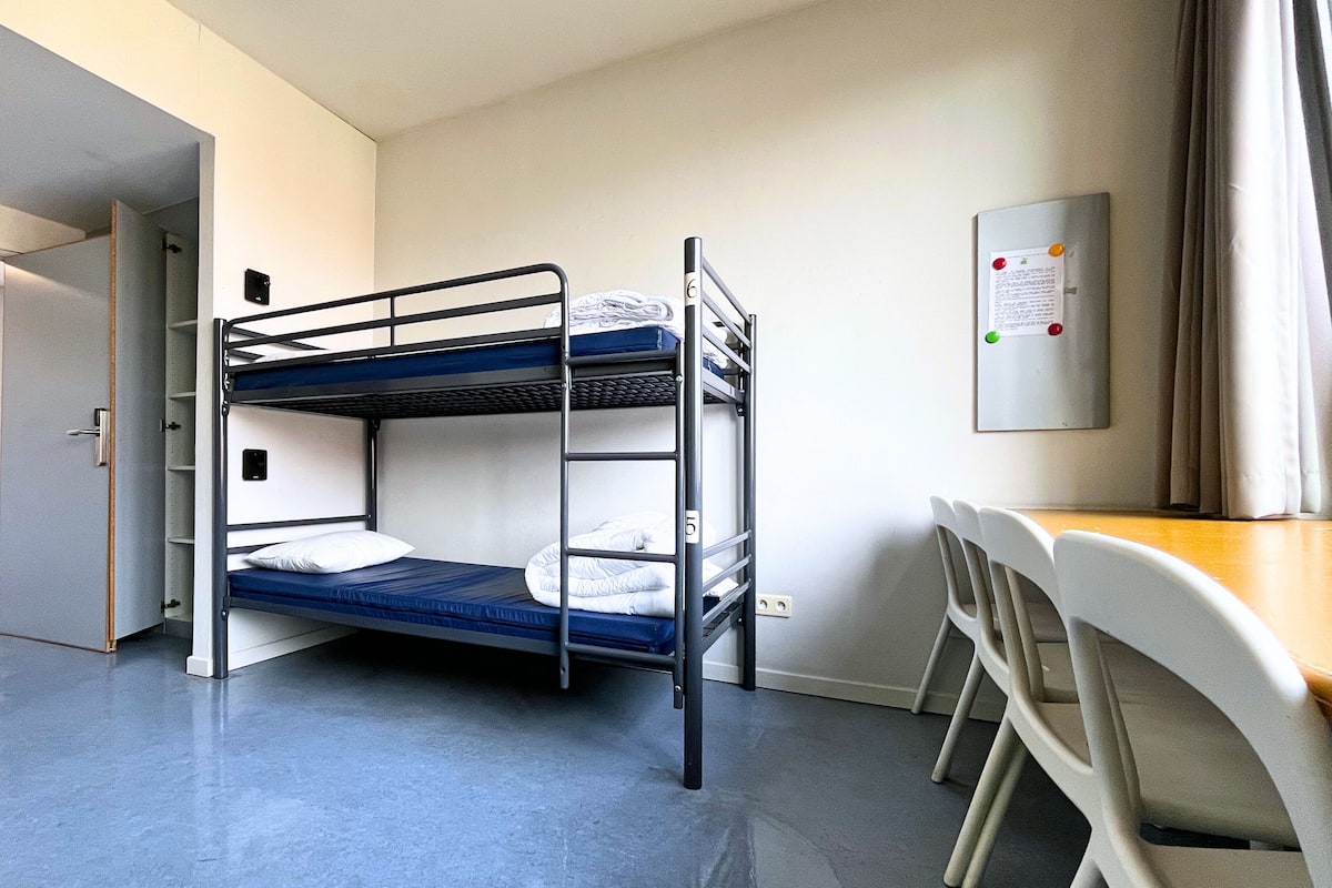 A bunk bed room with a desk and chairs, a door, and a wall-mounted notice board.