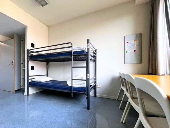 Habitación con literas, escritorio, sillas, una puerta y un tablón de anuncios fijado a la pared.