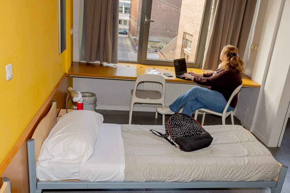 A single bed with a white mattress and a black and white patterned bag on it. A person is sitting on a white chair at a small table with a laptop.