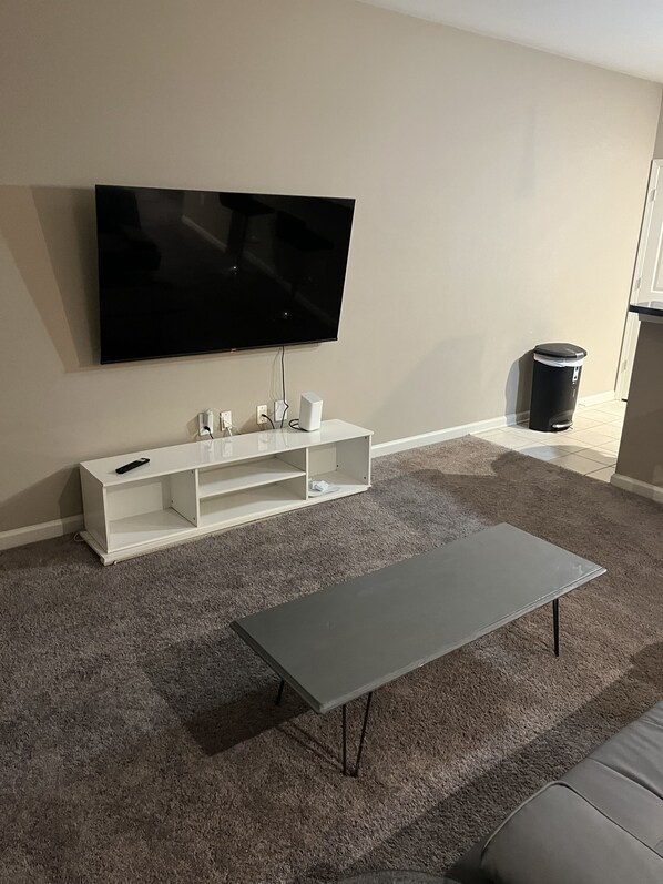 A living room with a flat-screen TV mounted on the wall, a white entertainment unit below, a grey coffee table, and a grey sofa. - Spacious 1 bedroom apartment in North ATL (Atlanta)