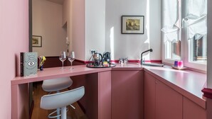 Una kitchenette rosa con una cafetera, dos copas de vino y un cuadro en la pared.