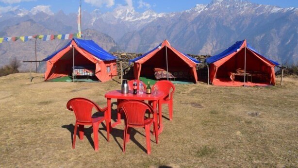 Three red tents with blue roofs, a table, and two red chairs in a grassy area with a mountainous backdrop. - Grand Auli Camp (Joshimath)