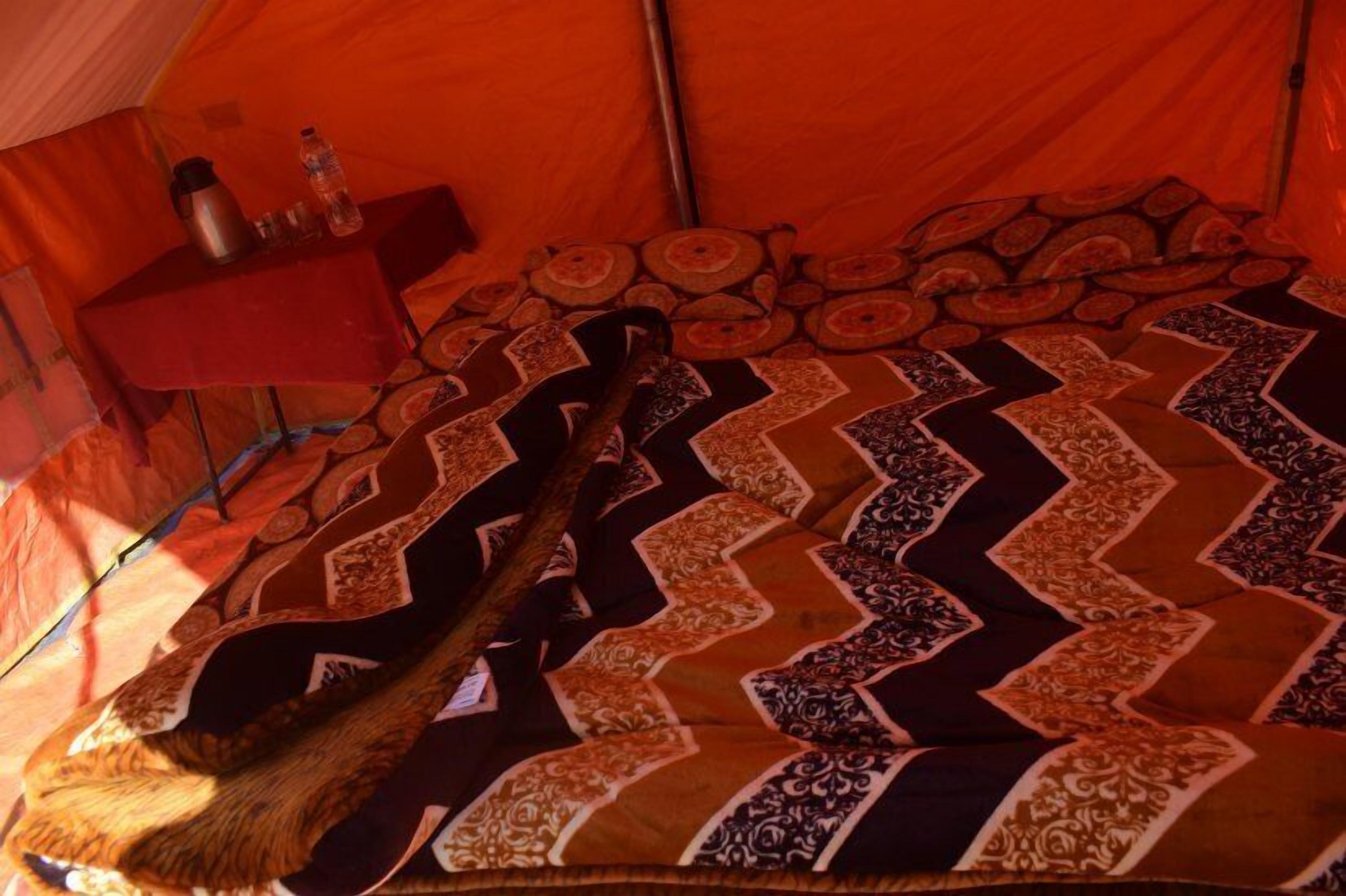 Interior de una tienda de campaña con una colcha estampada, una mesa roja con una botella de agua y un termo, y una zona para dormir.