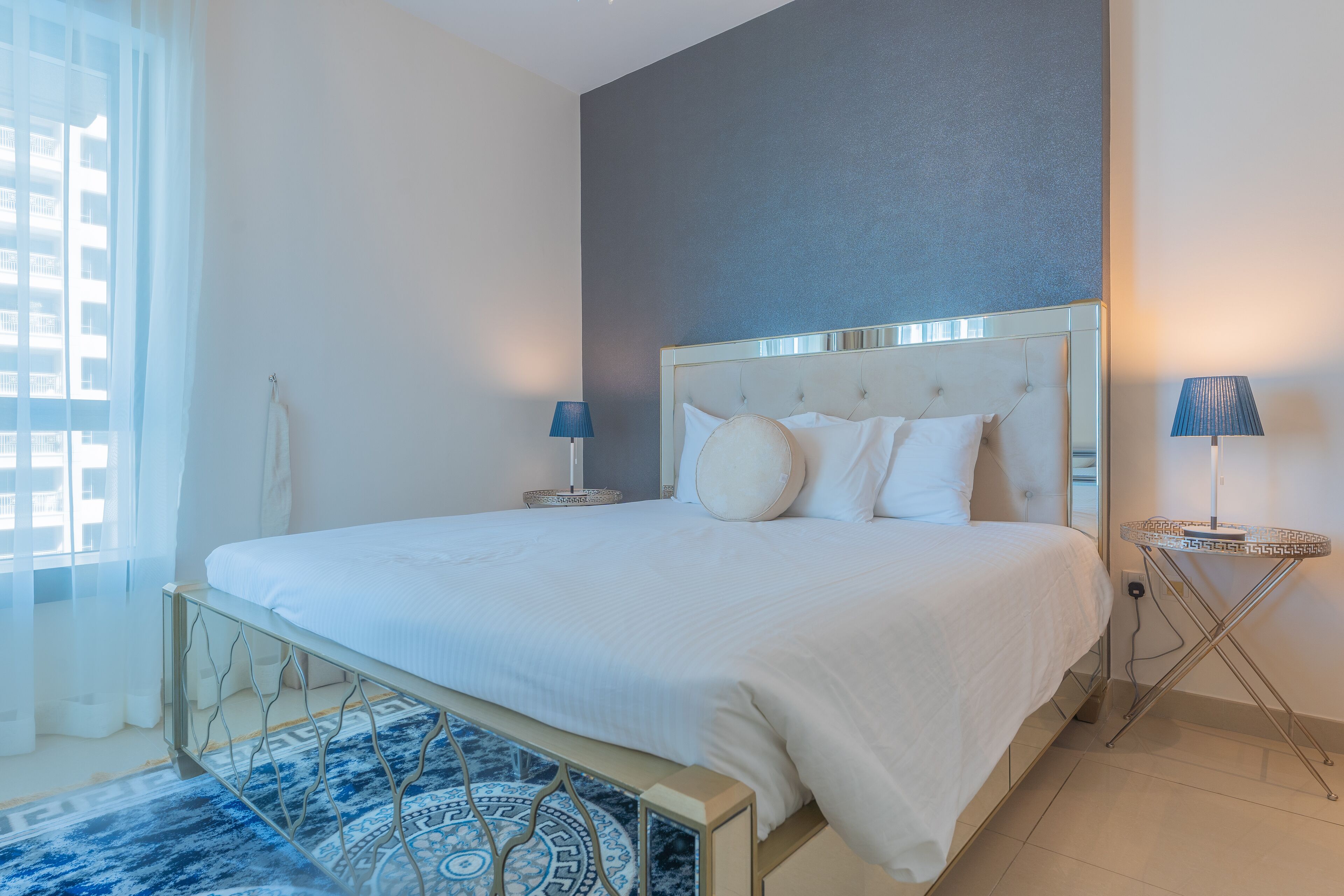 A modern hotel room with a large bed, two bedside tables with lamps, a patterned rug, and a view of the city through the window.