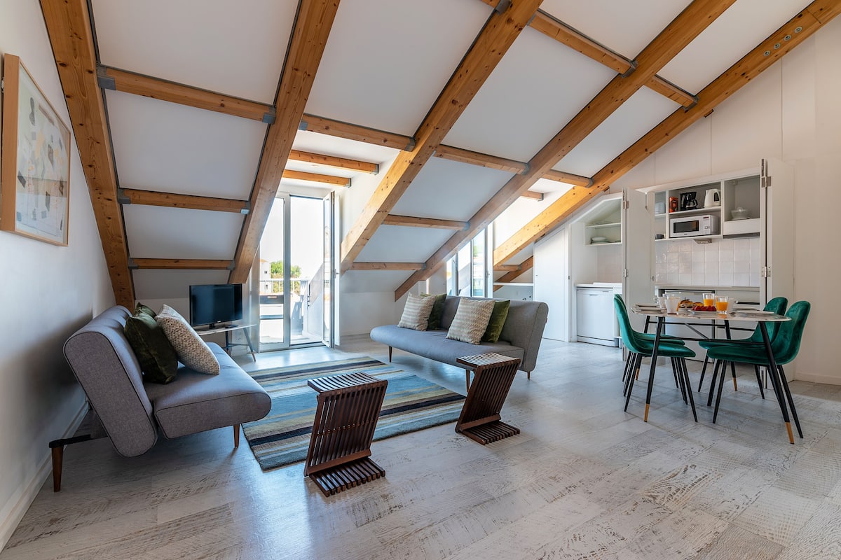 A modern loft-style living area with a high ceiling, exposed wooden beams, a grey sofa, a dining table with chairs, and a kitchen area with cabinets and appliances.