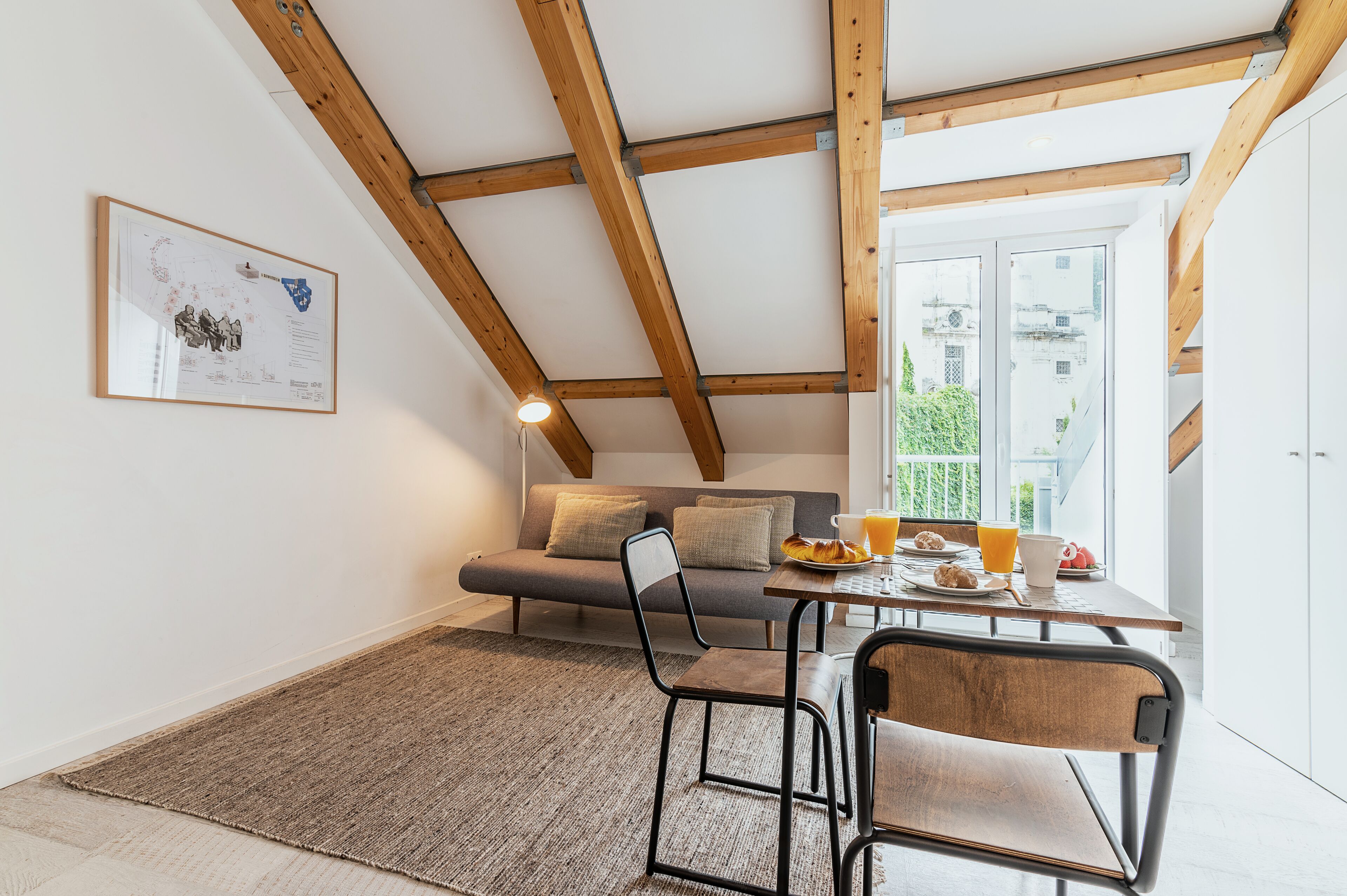 Ein modernes Schlafzimmer im Loft-Stil mit freiliegenden Holzbalken, einem großen Fenster, einer gemütlichen Couch und einer Frühstücksecke an einem Holztisch.