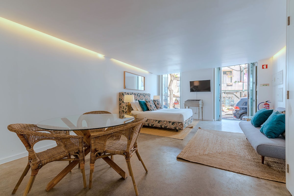 A modern, well-lit room with a glass dining table, wicker chairs, a sofa, and a bed with a view of the street.